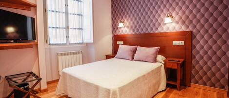 Une chambre à coucher comprenant un lit, des tables de chevet, un téléviseur fixé au mur, un radiateur et une fenêtre avec des rideaux.