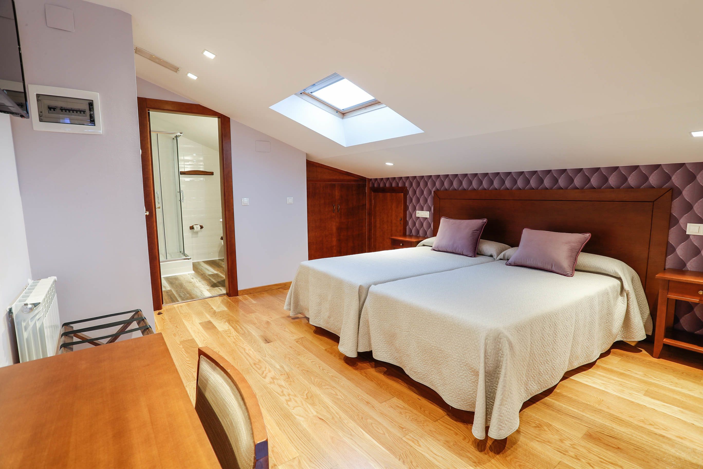 Un dormitorio con dos camas, un tragaluz, piso de madera y un baño visible a través de una puerta abierta.