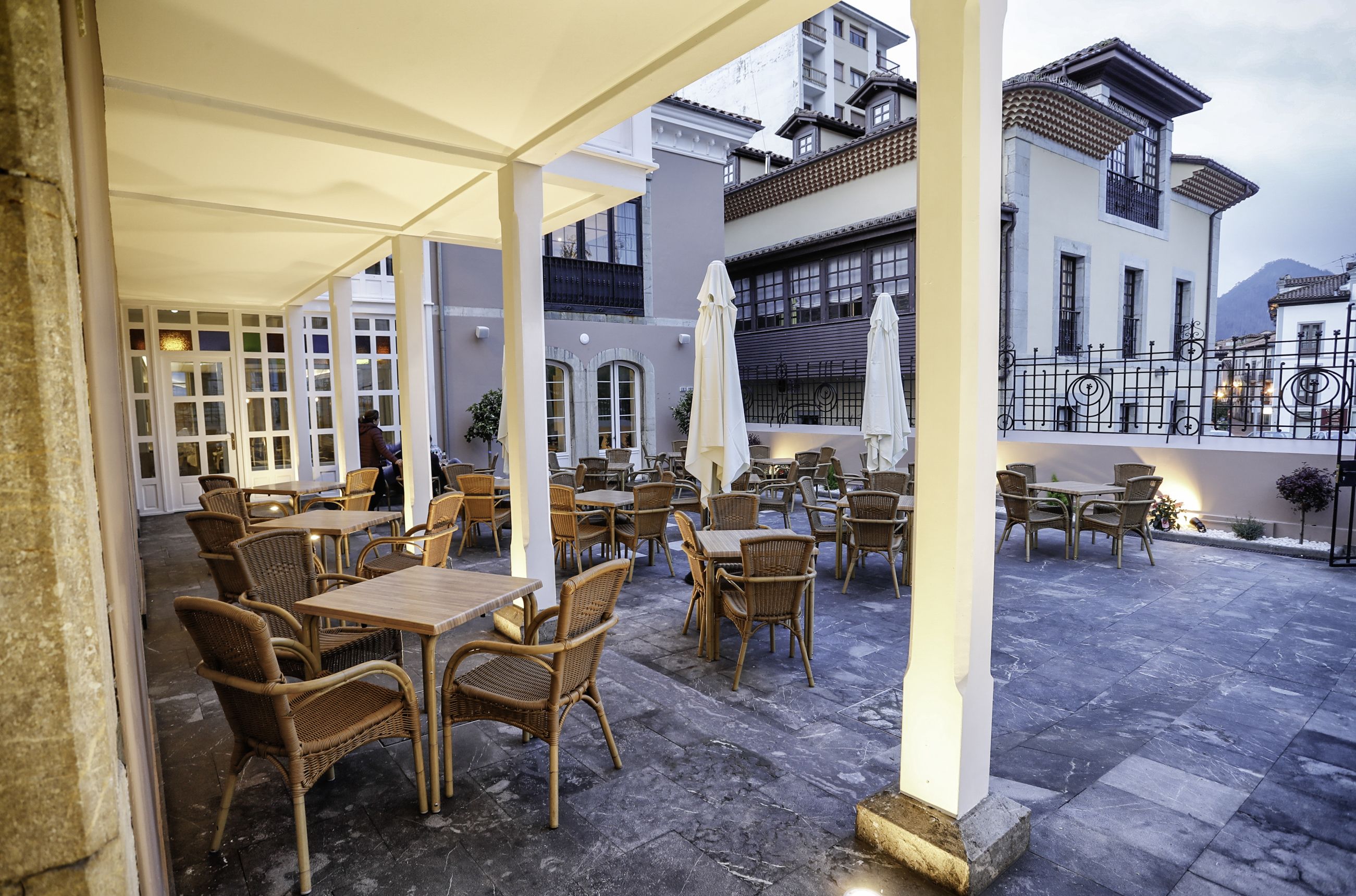 An outdoor cafe area with wicker furniture and umbrellas, set against a backdrop of traditional buildings and a mountain.