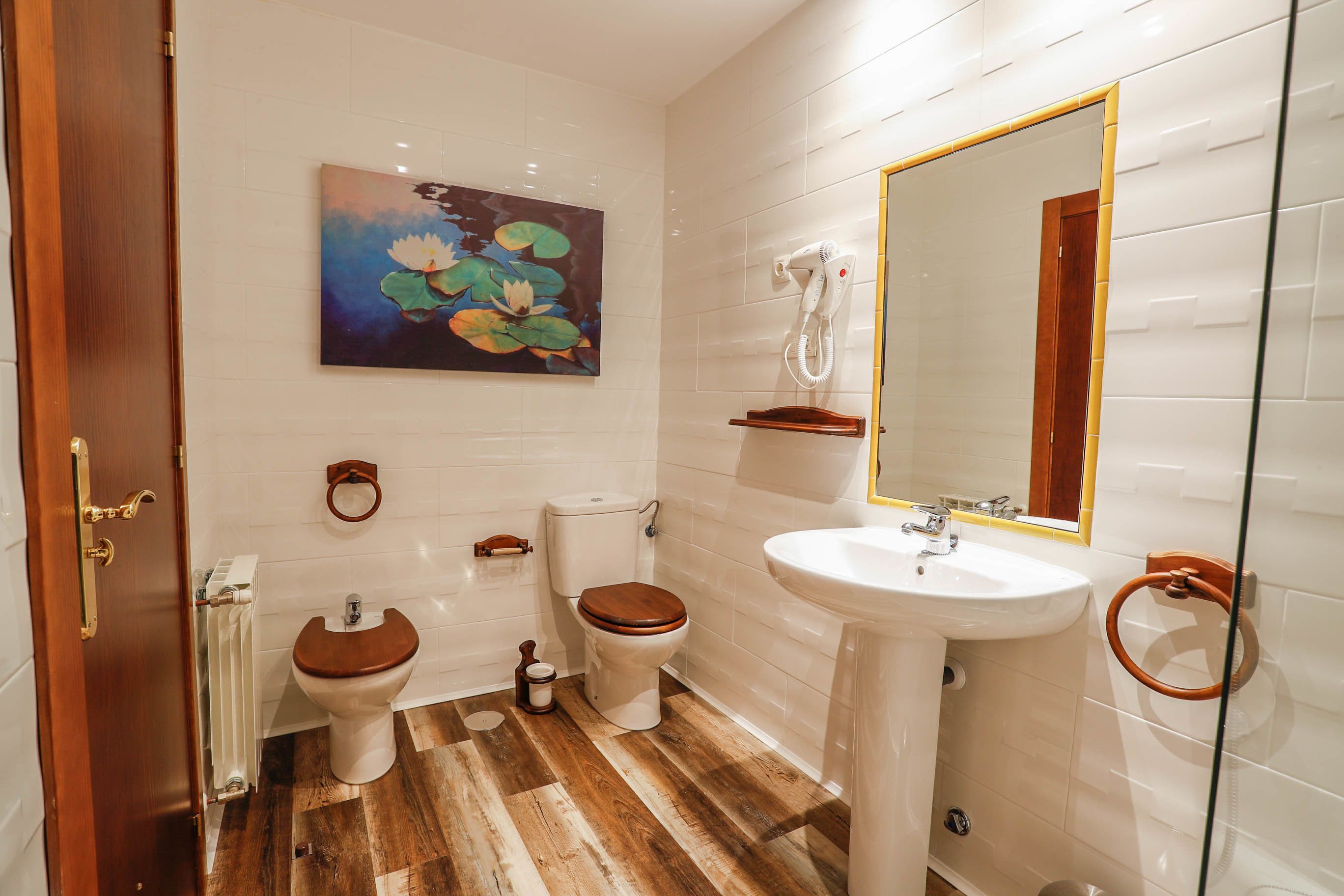 Een badkamer met een houten vloer, witte tegels, een toilet, een bidet, een wastafel en een spiegel.