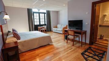 A hotel room with a bed, a desk with a TV, a chair, a small table, and a window with curtains.