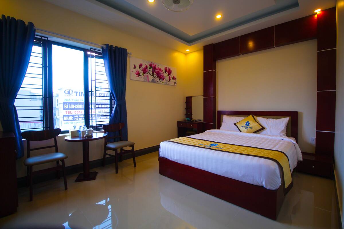 Habitación de hotel con una cama grande, una pequeña mesa con sillas, una ventana con cortinas y un cuadro en la pared.