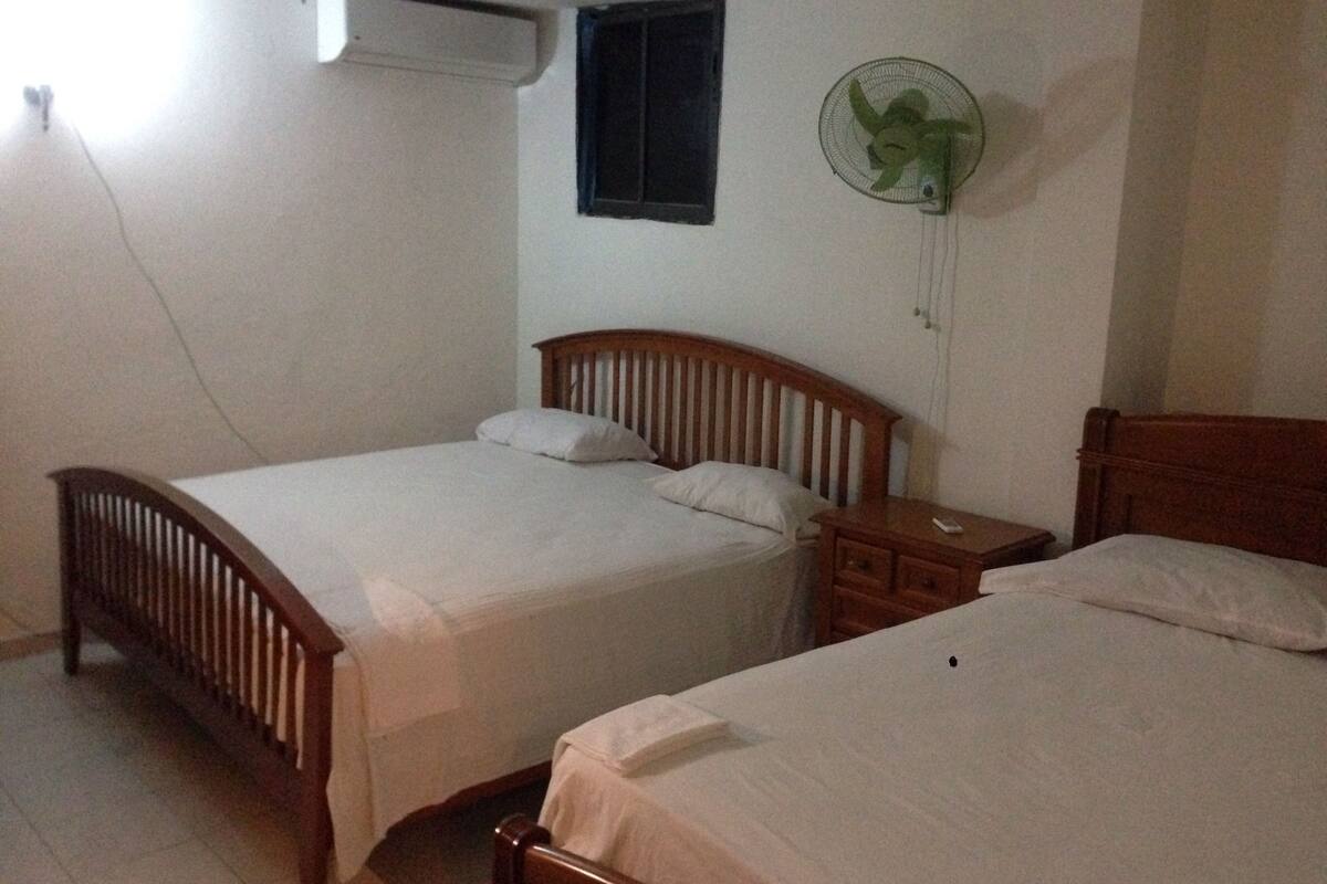 A bedroom with two wooden beds, a small bedside table, a wall-mounted fan, and an air conditioning unit.