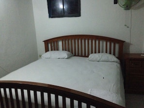 Quarto individual com cama de solteiro, cabeceira de madeira, roupa de cama branca e mesa de cabeceira pequena com abajur.