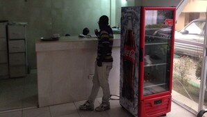 Un homme se tient dans une pièce où l’on aperçoit un réfrigérateur Coca-Cola, un bureau avec un ordinateur et une fenêtre donnant sur une voiture garée.