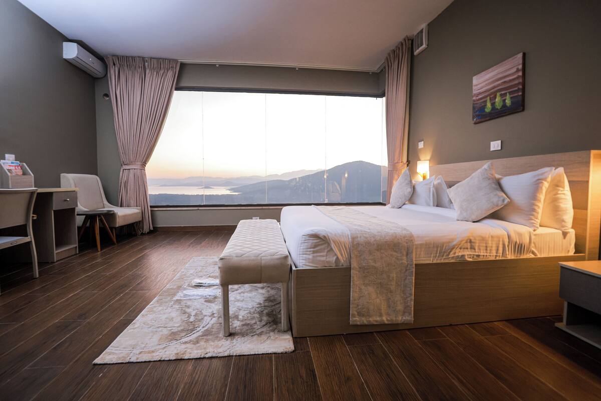 A hotel room with a large bed, a desk, a chair, a view of the mountains, and a painting on the wall.