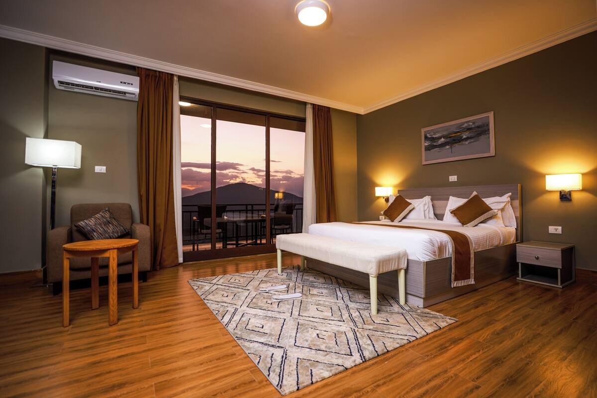 A hotel room with a bed, bedside tables, a chair, a lamp, an air conditioner, and a view of the sunset through the window.