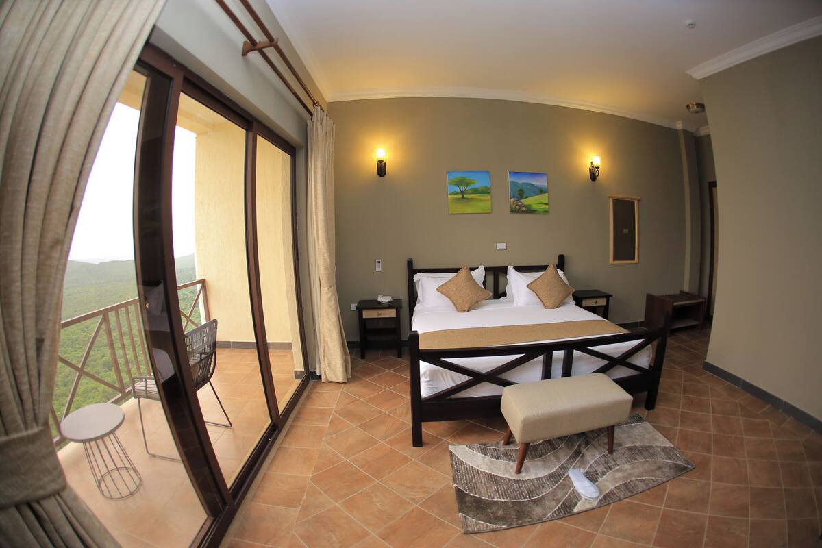 A bedroom with a large bed, a small table, a chair, a nightstand, and a view of the outdoors.