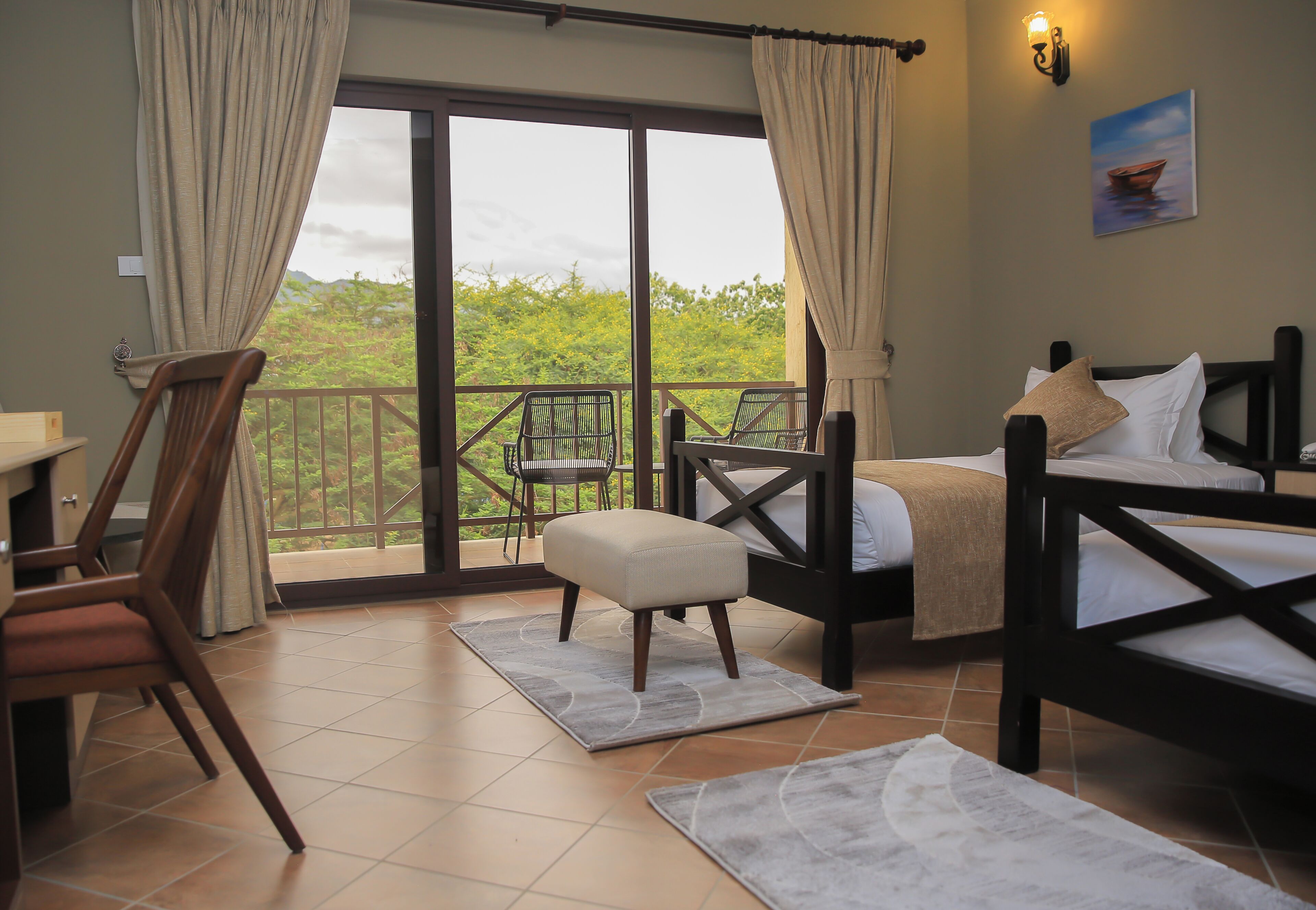 A room with a bed, a chair, a table, and a balcony with a view of greenery.