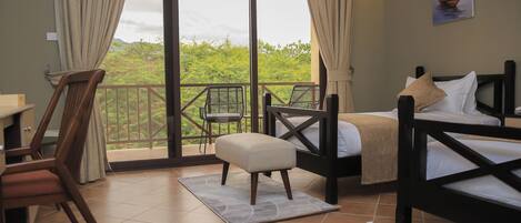 A room with a bed, a chair, a table, and a balcony with a view of greenery.