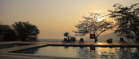 A swimming pool with lounge chairs and a tree in the background during sunset.