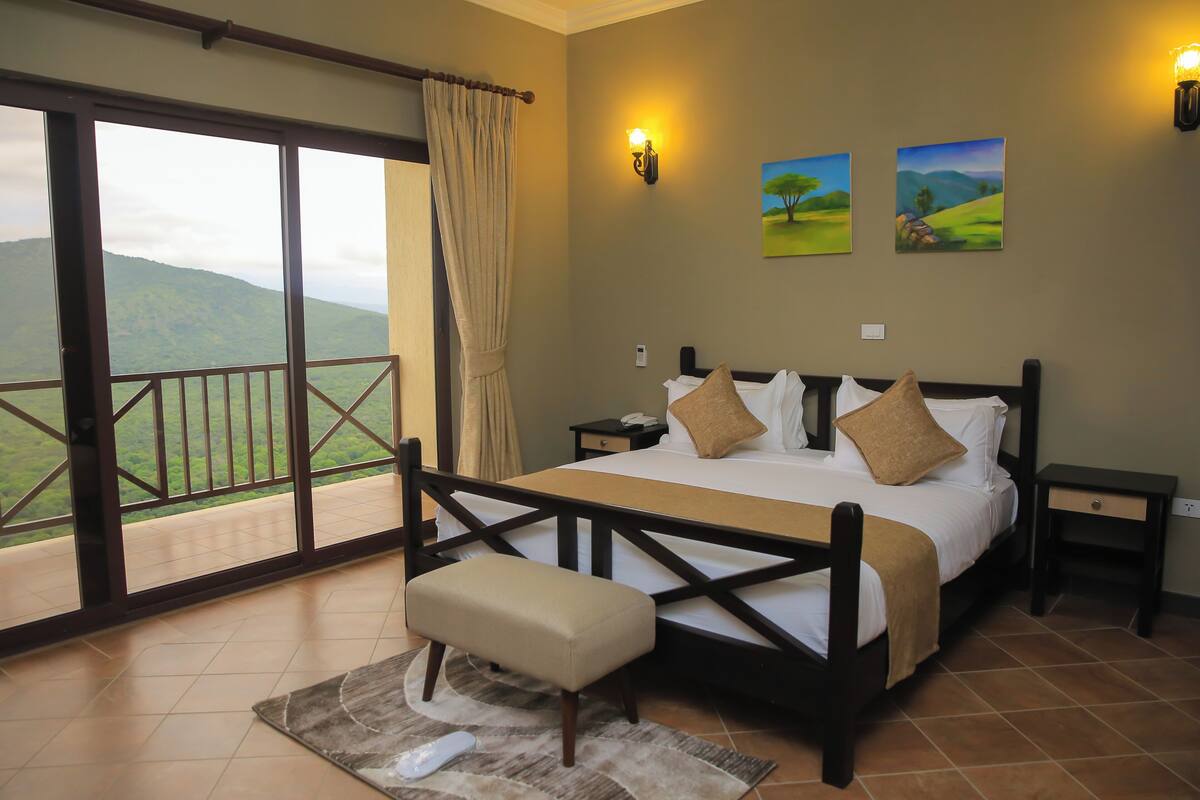 A bedroom with a large bed, a bench, a nightstand, and a view of green hills.
