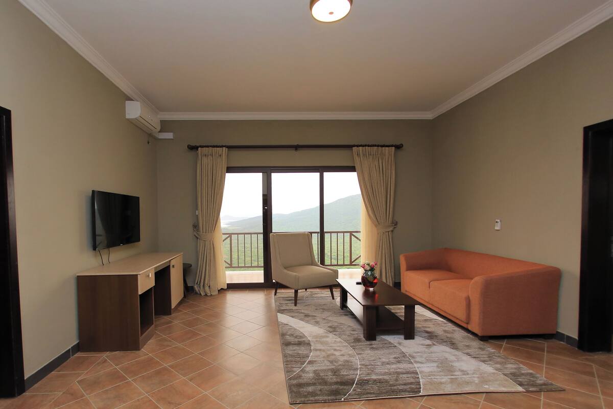 A living room with a flat-screen TV, a sofa, a chair, a coffee table, and a view of the outdoors through a sliding door.