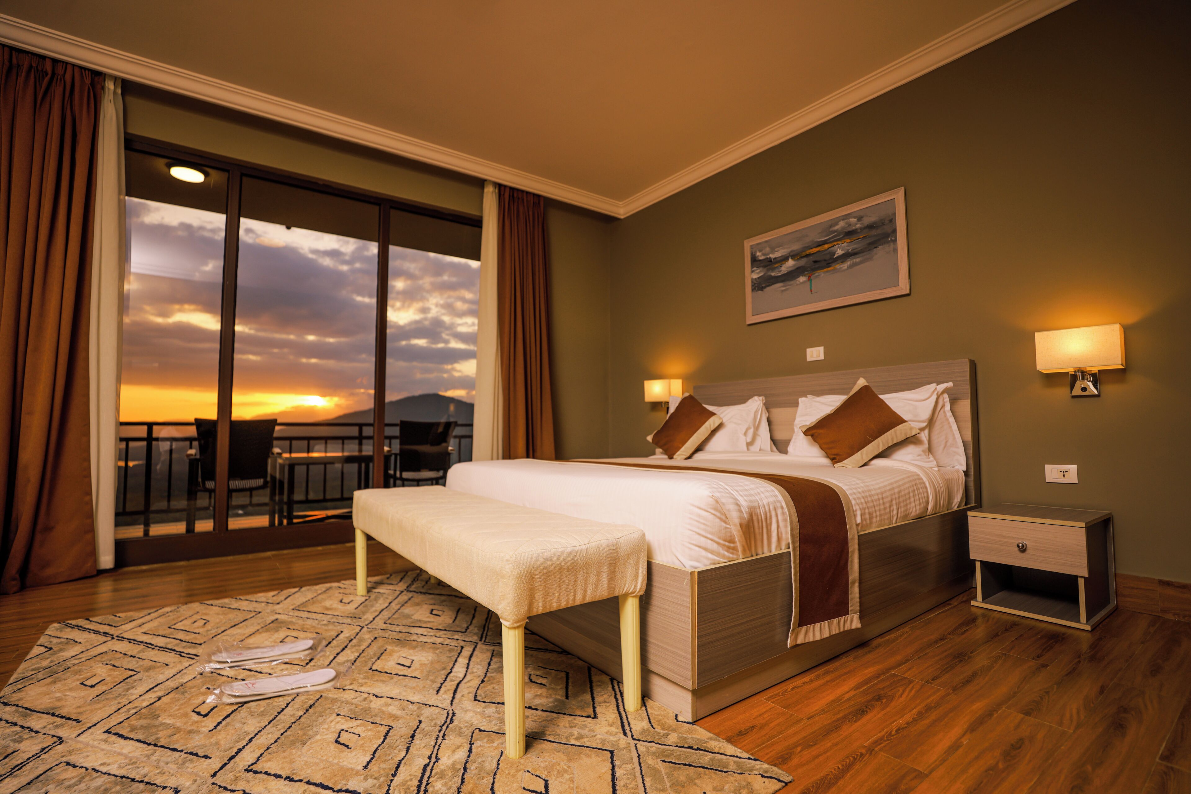 A hotel room with a bed, bedside tables, a bench, a view of the sunset, and a balcony.