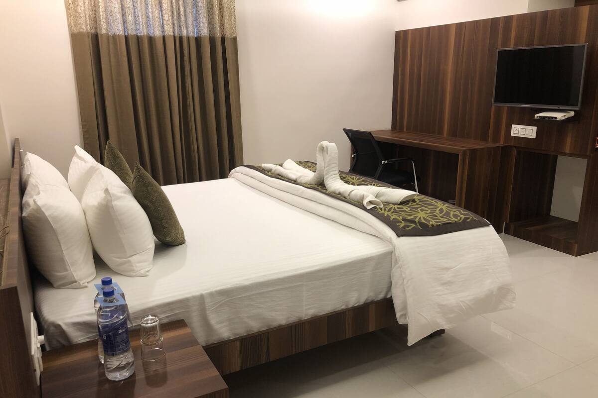 A hotel room with a neatly made bed, a bedside table with a water bottle, a TV mounted on the wall, and a wooden desk.