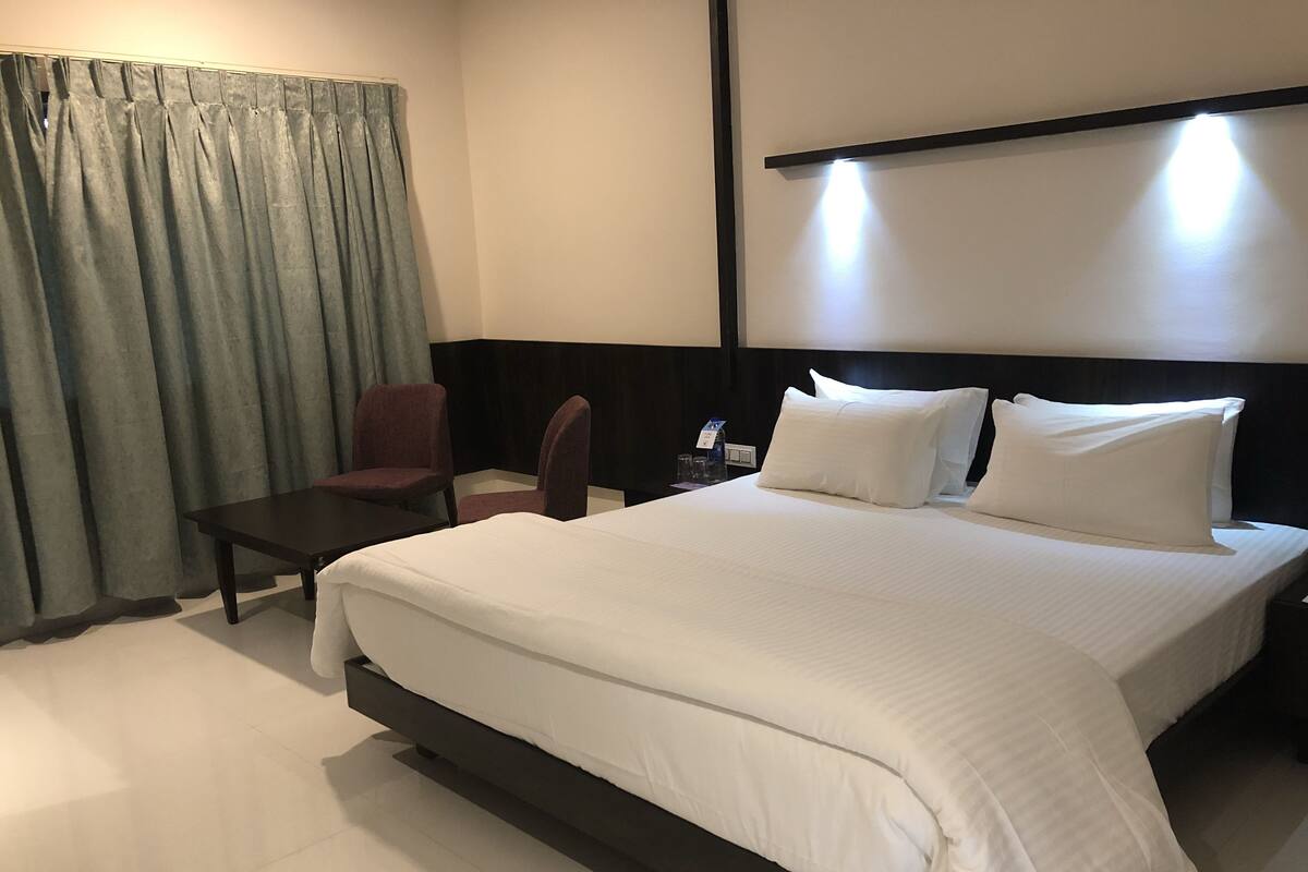 A hotel room with a large bed, a small table, two chairs, a nightstand, and a curtain.