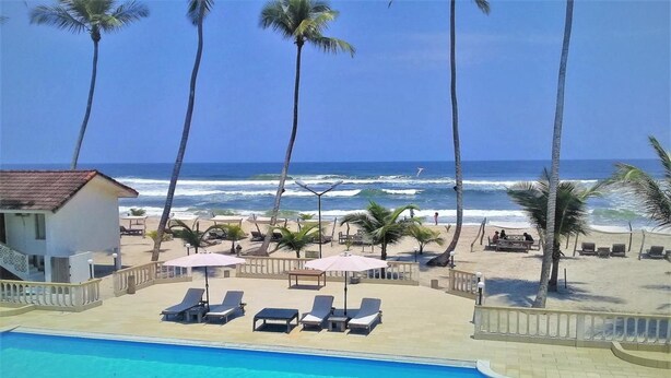 Zona de piscina con sillones de descanso y sombrillas, con vista a una playa con palmeras y al océano.