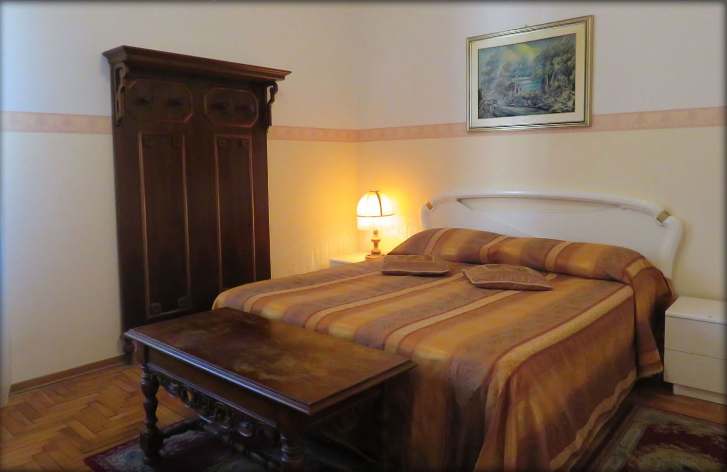 Un dormitorio con cama, mesita de noche, lámpara, armario de madera y un cuadro en la pared.