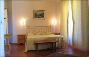 A bedroom with a bed, bedside tables, a bench, a painting on the wall, and a window with curtains. - Casalbergo (Siena)