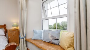 A bedroom with a wooden bed, a nightstand with a lamp, a window seat with cushions, and a view of greenery outside. - Treweens Bed & Breakfast (Wadebridge)