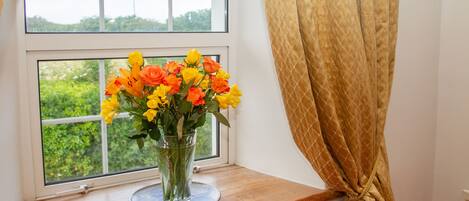 A window with a view of greenery, a vase with flowers, and a curtain.