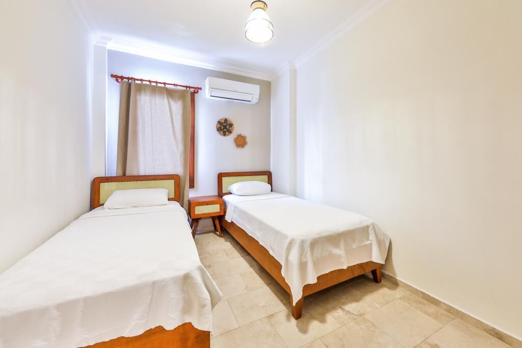 A room with two single beds, a small bedside table with a lamp, an air conditioning unit, and a window with curtains.