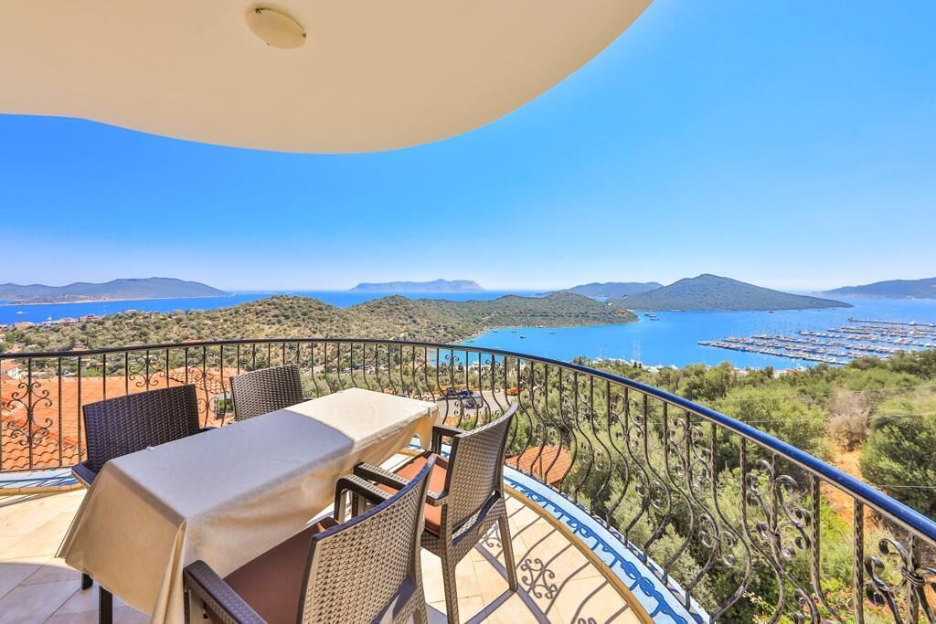 Un balcon avec une table et des chaises donnant sur un paysage pittoresque, avec une étendue d’eau et des collines.