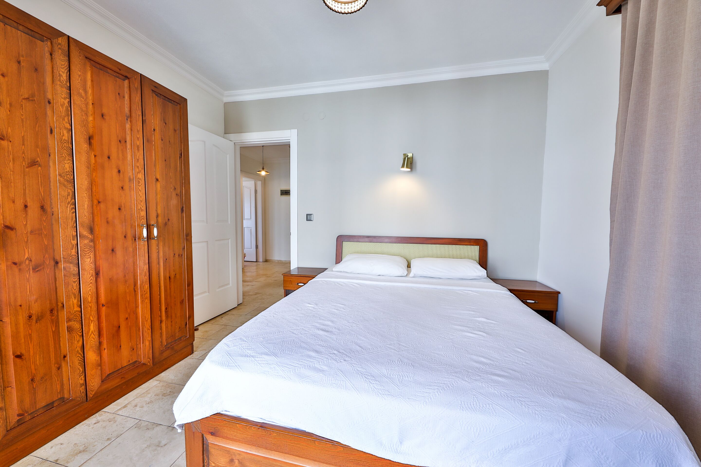 A bedroom with a large wooden wardrobe, a bed with white bedding, a nightstand, and a wall-mounted light.