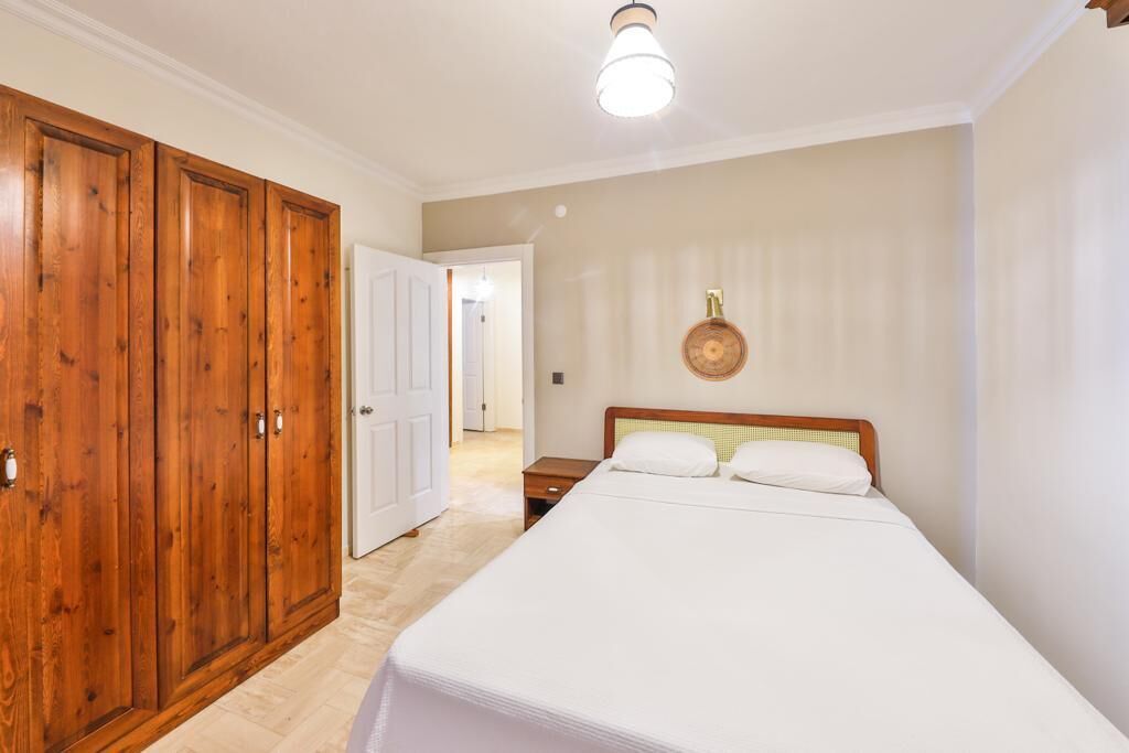 A bedroom with a wooden wardrobe, a bed with white bedding, and a small bedside table.