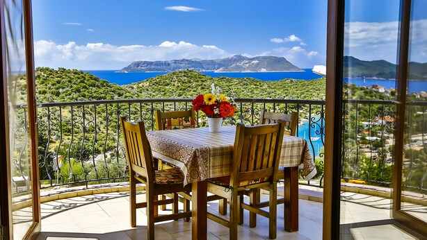 Un balcon avec une table et des chaises, un vase de fleurs et une vue panoramique sur la mer et les montagnes.