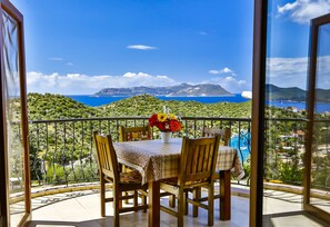 Un balcó amb taula i caires, un vas de flors i una vista panoràmica del mar i les muntanyes.