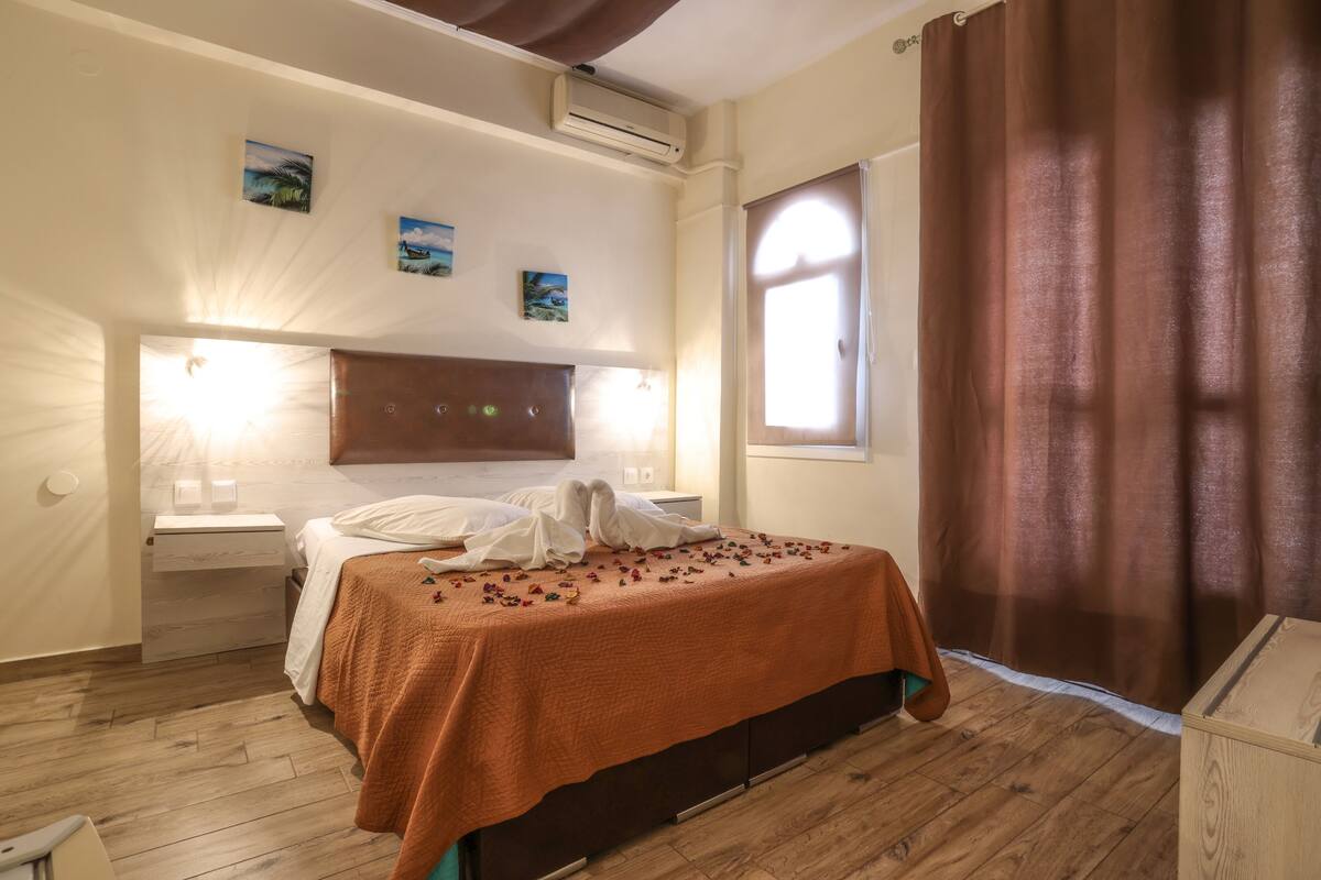 A bedroom with a wooden floor, a bed with a brown bedspread, a nightstand, a wall-mounted air conditioner, and a window with brown curtains.