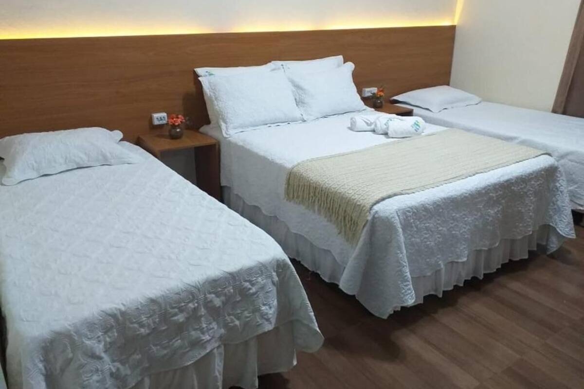 Ein Hotelzimmer mit zwei Betten, jeweils mit weißen Bettwäsche und Spitzenüberzügen. Es gibt ein hölzernes Kopfteil, einen Nachttisch mit einer Lampe sowie einen kleinen Tisch mit einer Blumenvase.