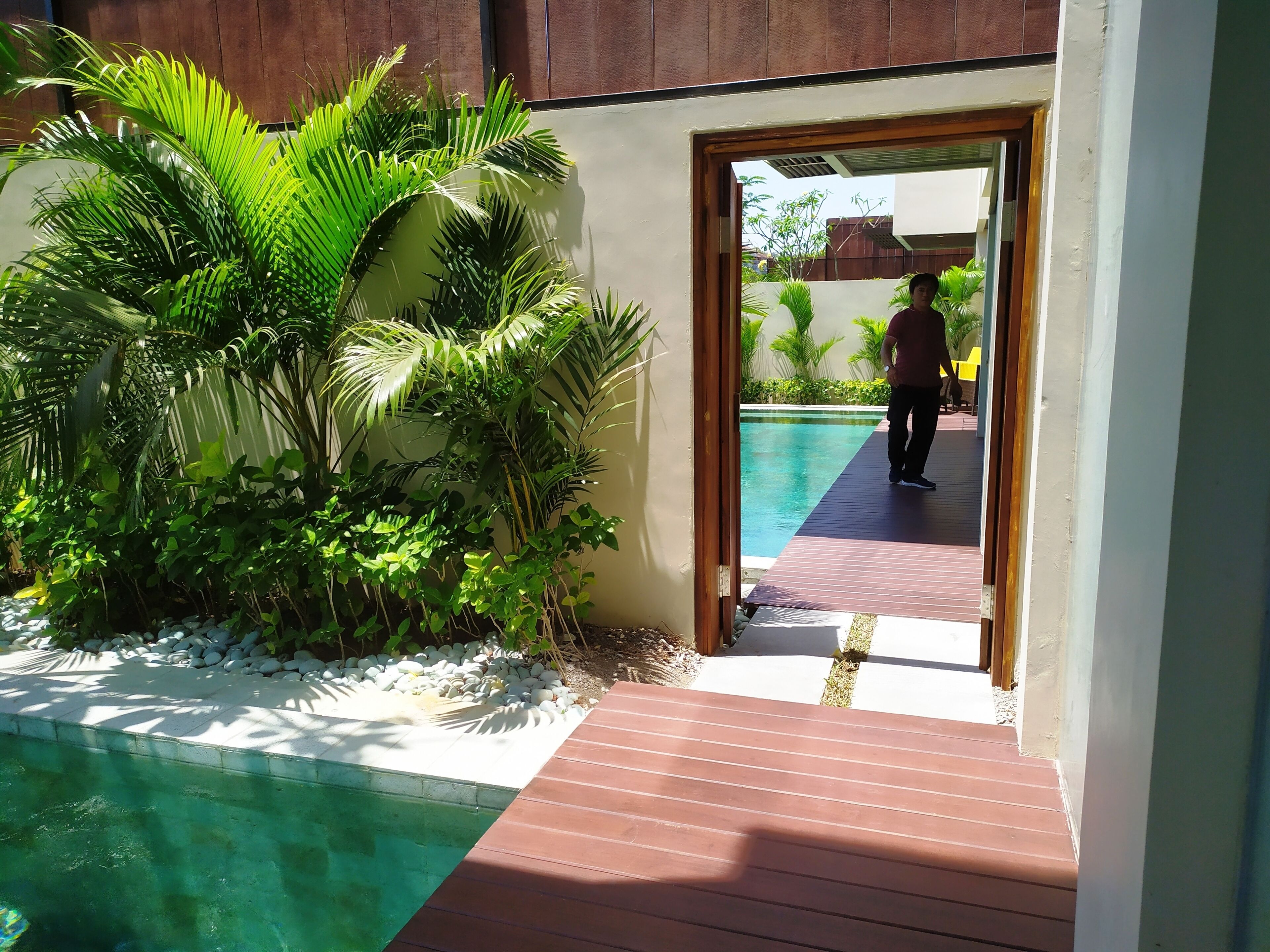 A pool area with a wooden deck leading to a pool, surrounded by lush greenery and a clear blue pool.