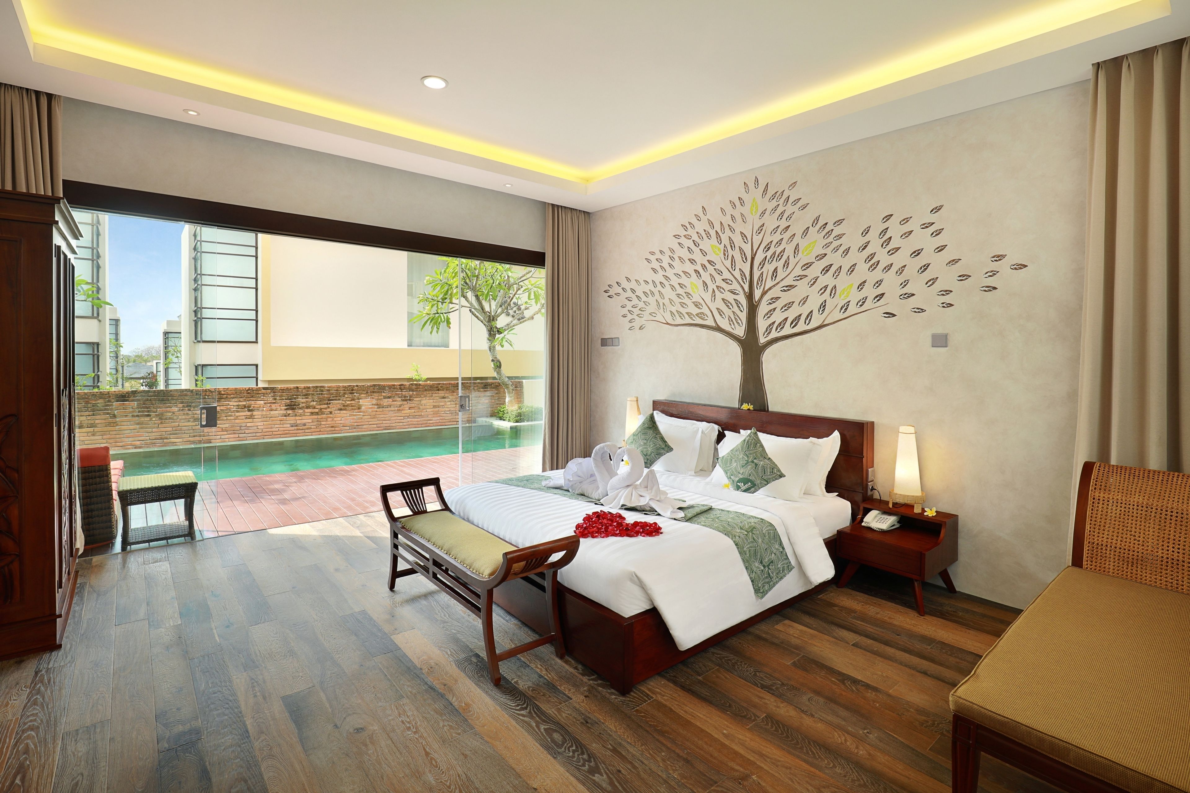 A modern hotel room with a large bed, a bedside table with a lamp, a small table with a chair, and a view of a swimming pool and buildings outside.