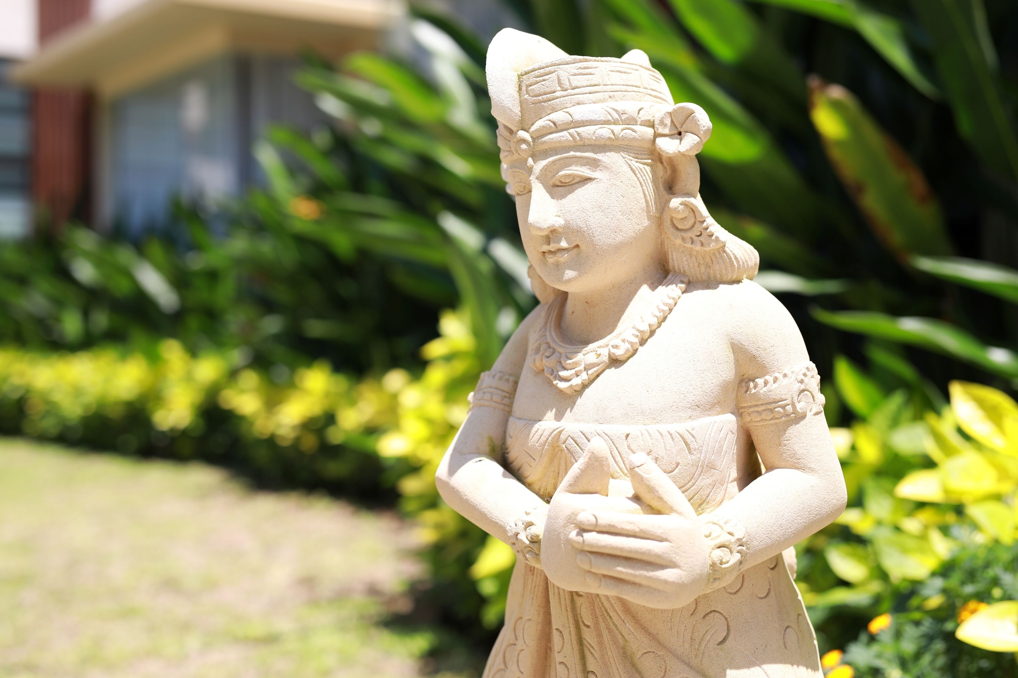 Una estatua de una mujer con traje tradicional, ubicada en un jardín con abundante vegetación y un edificio al fondo.