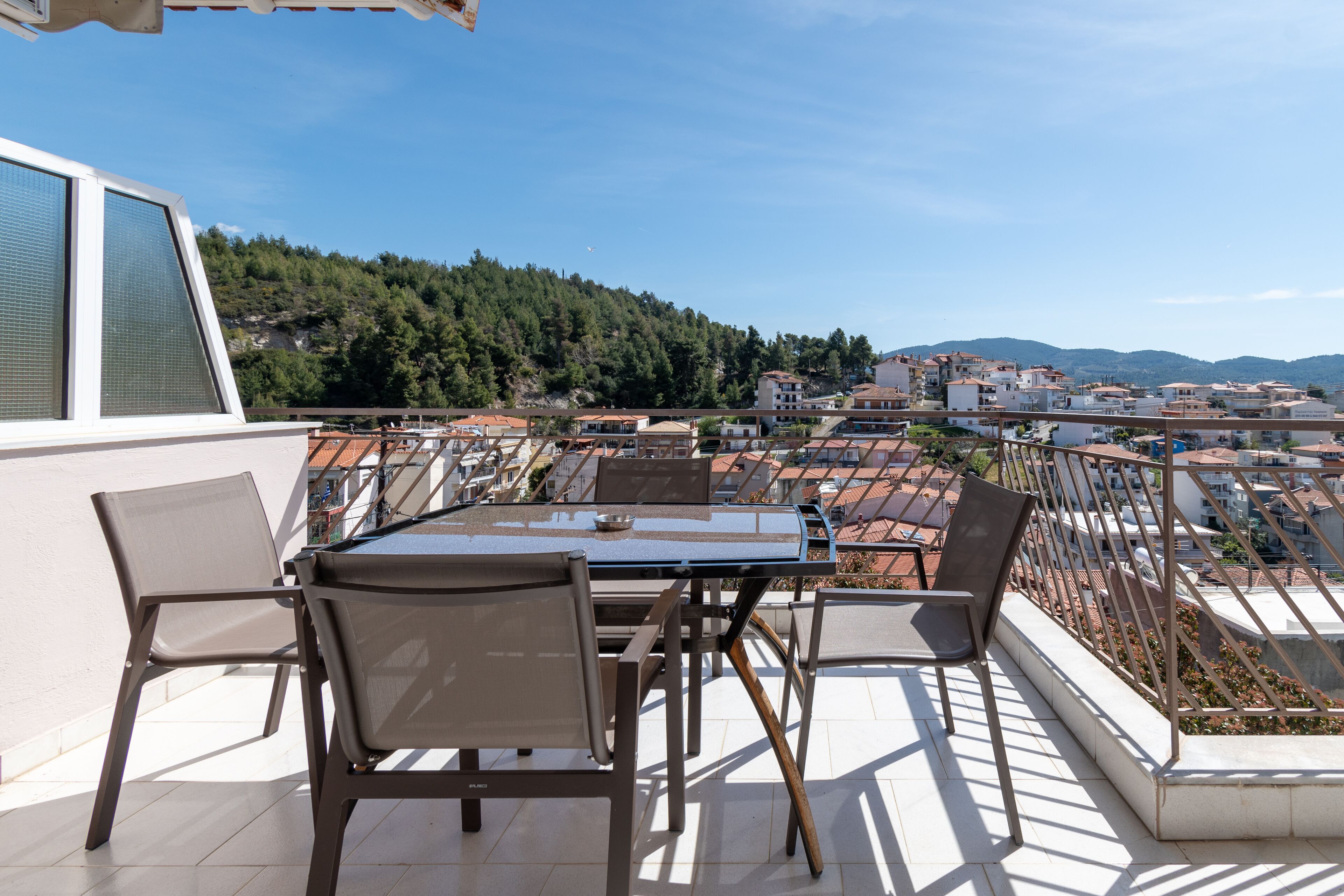 En terrasse med et bord og stole, med udsigt over et boligområde med huse og en bakke i baggrunden.