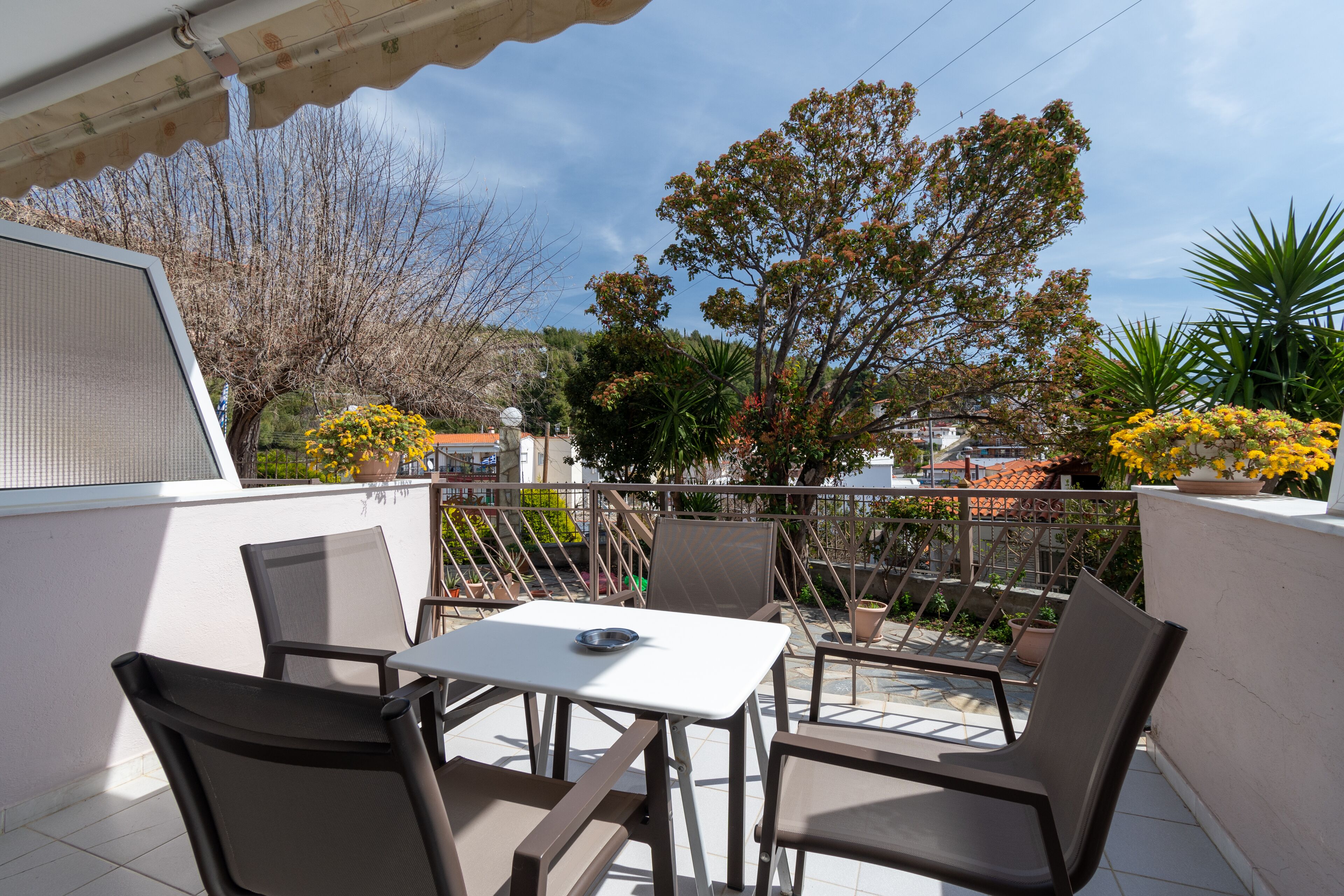 A balcony with a table and chairs, surrounded by potted plants and a view of buildings and trees.
