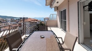 Varanda com mesa e cadeiras, vista para área costeira com edifícios e um corpo de água.