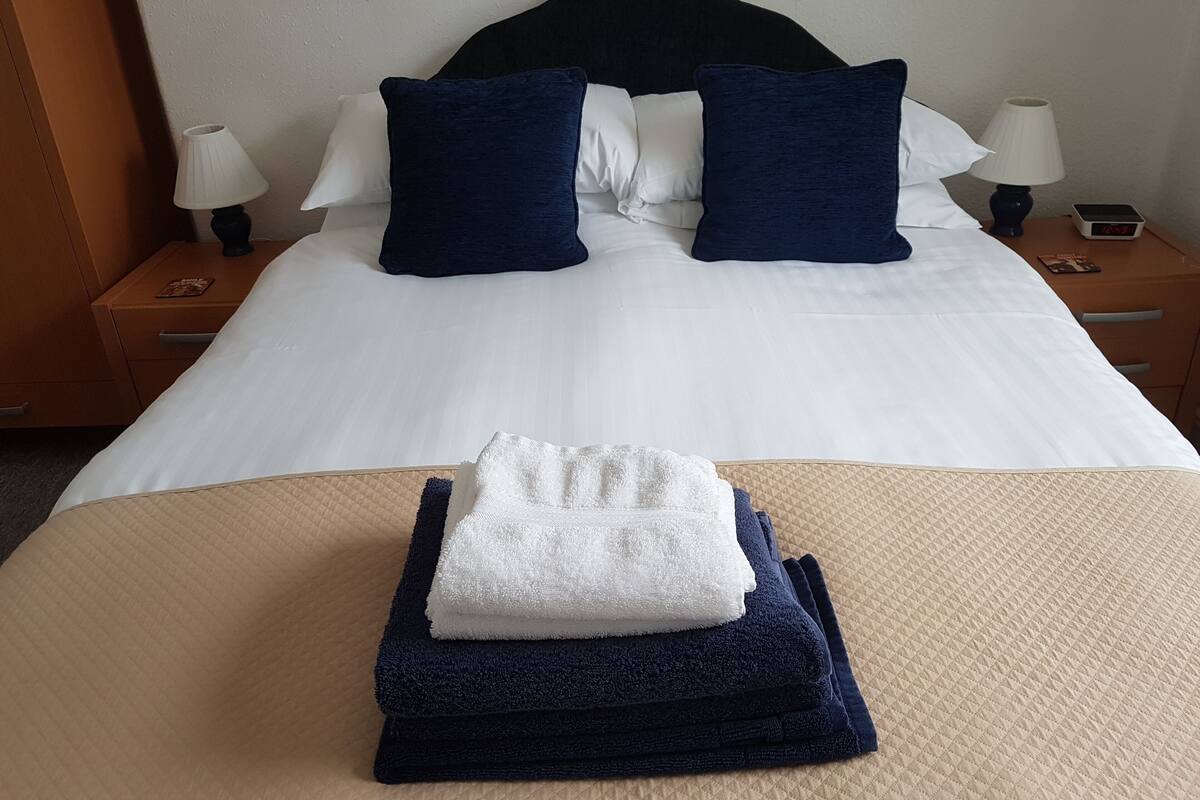 Una cama bien hecha con edredón blanco, dos almohadas azul marino y una toalla blanca doblada sobre un corredor beige.