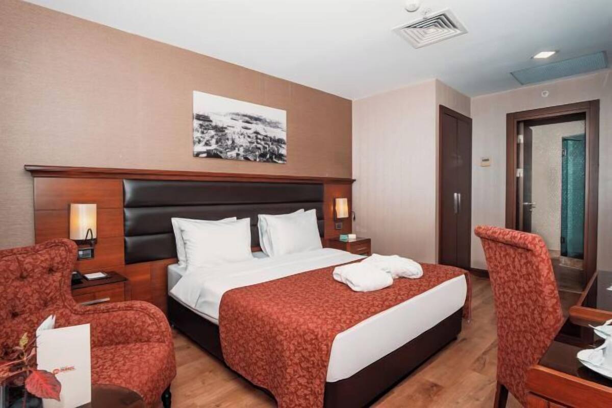 A hotel room with a large bed, two bedside tables, a nightstand, a chair, a desk, and a framed picture on the wall.