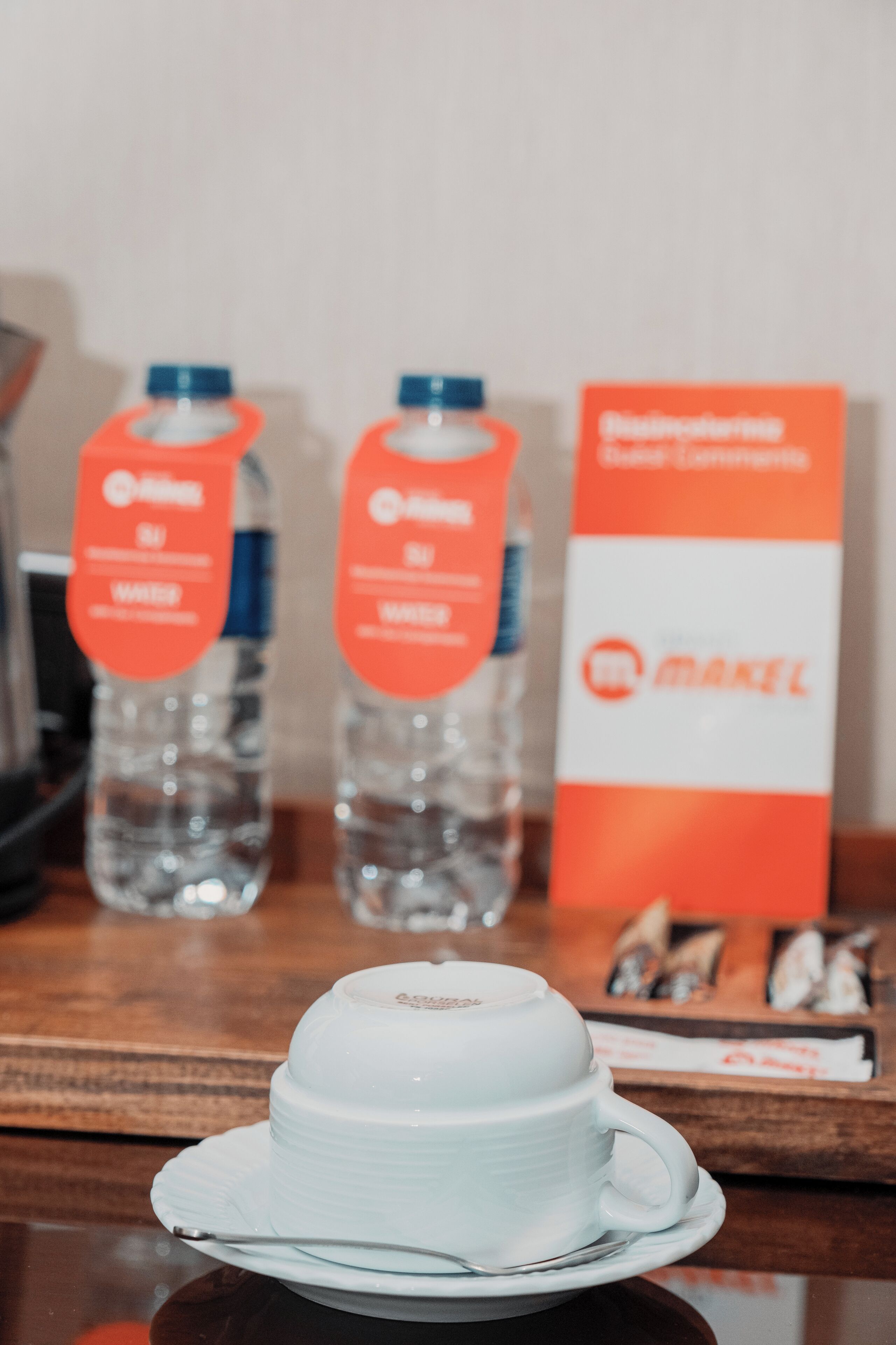 A white cup on a saucer, two water bottles, and a box labeled "MAKEL" on a wooden table.