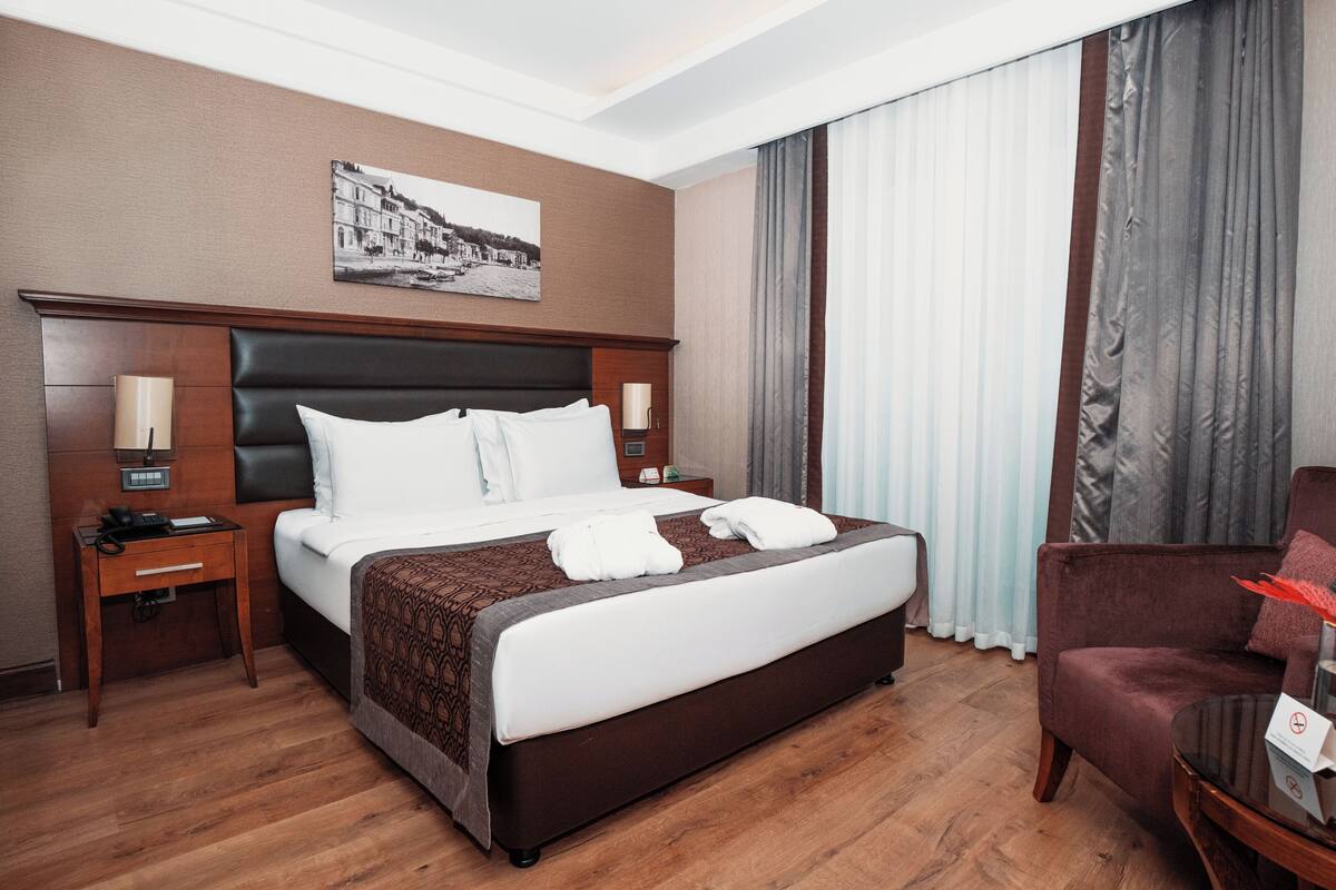 A hotel room with a large bed, a wooden headboard, a nightstand with a lamp, a chair, and a painting on the wall.