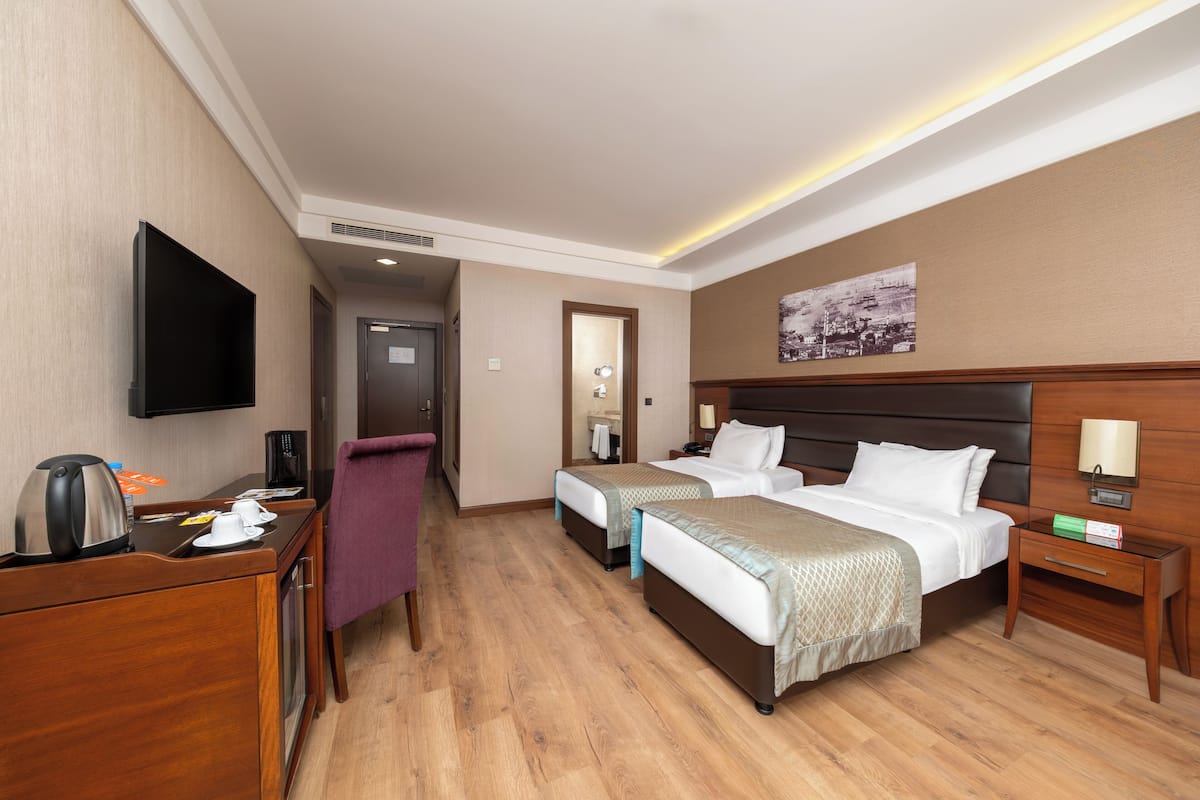 A hotel room with two beds, a desk with a kettle, a chair, a TV, and a painting on the wall.