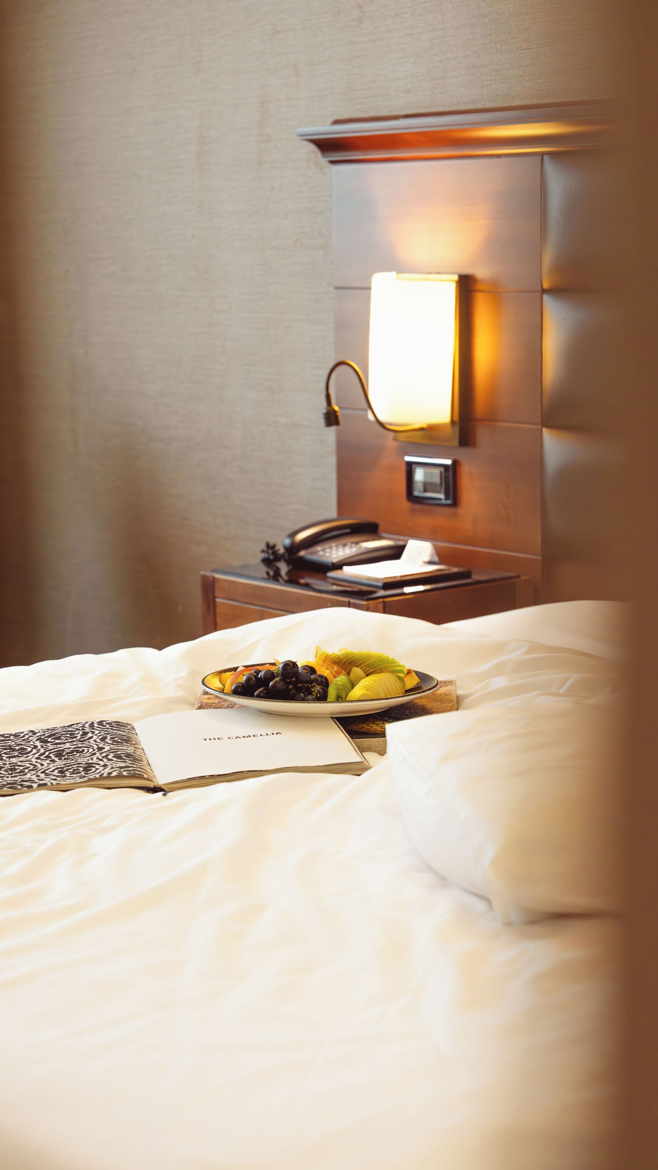 A hotel room with a neatly made bed, a bedside table with a lamp, a telephone, and a plate of fruit.