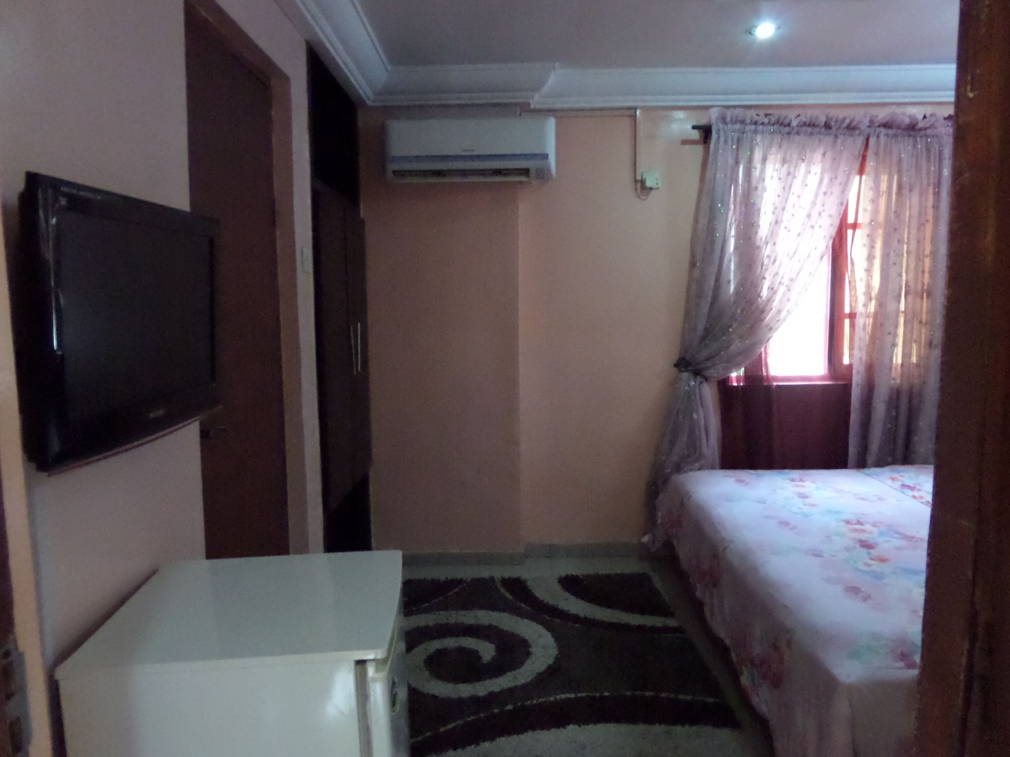 A small, dimly lit room with a bed, a TV mounted on the wall, and a window with sheer curtains.