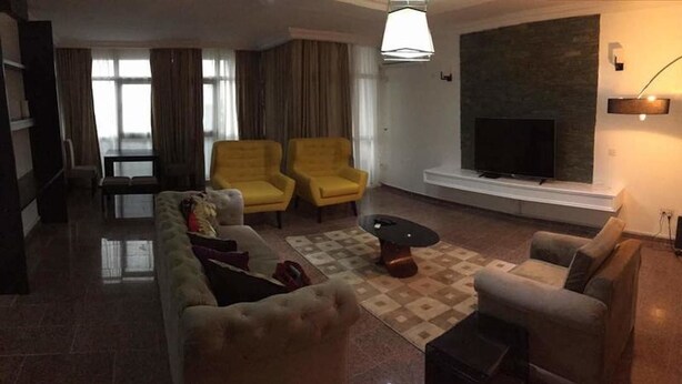 A living room with a sofa, two yellow armchairs, a coffee table, and a TV mounted on the wall. - Koza Apartment (Lagos)