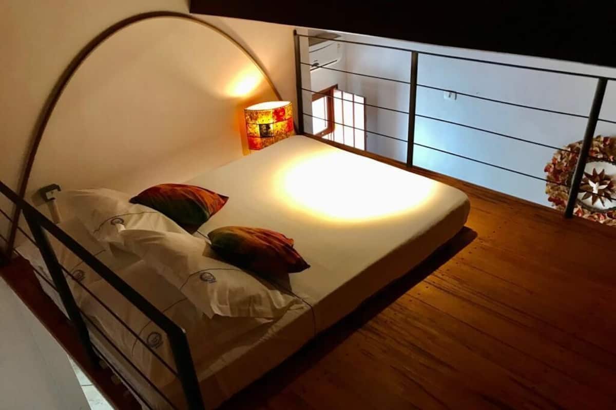 Uma cama pequena com cabeceira curva, uma luminária de cabeceira e vista para uma varanda com grade e uma coroa decorativa.