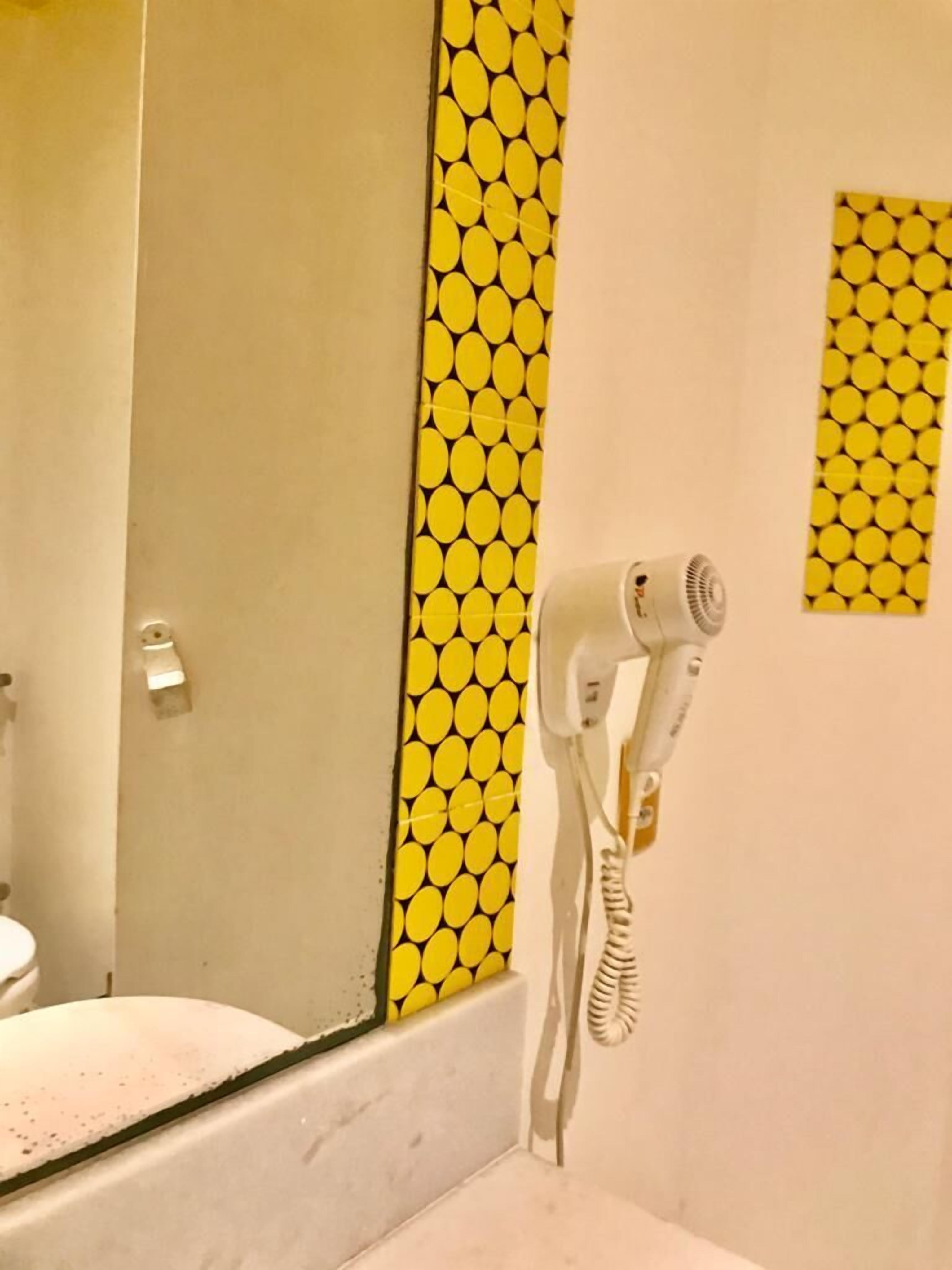 Un baño con pared amarilla estampada, un secador de pelo blanco montado en la pared y un espejo sobre un lavamanos.