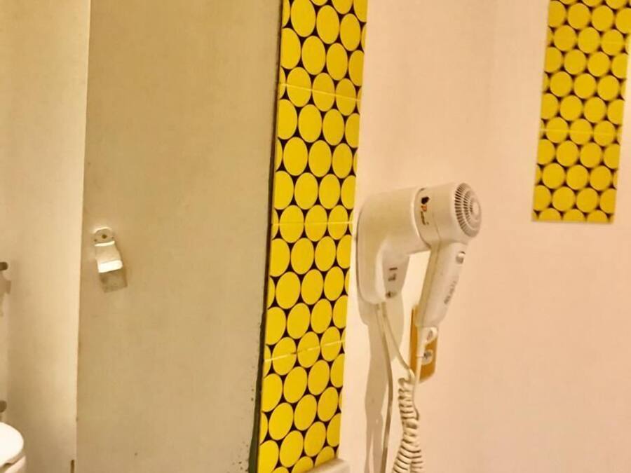 Un baño con pared amarilla estampada, un secador de pelo blanco montado en la pared y un espejo sobre un lavamanos.